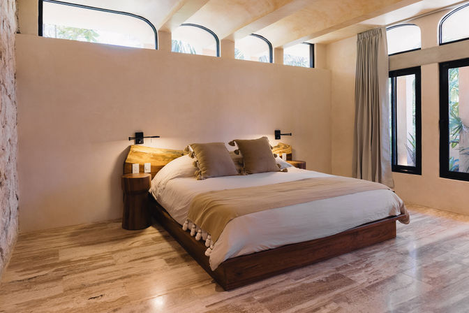 Cozy sunlit minimalist bedroom with low wooden platform bed, neutral linens, arched clerestory windows, and stone tile floor.