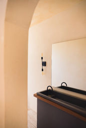 Sunlit minimalist bathroom interior in warm beige tones with an arched plaster doorway, large rectangular mirror, black double basin sink with curved faucets, wood-trimmed counter and exposed-bulb wall sconce.