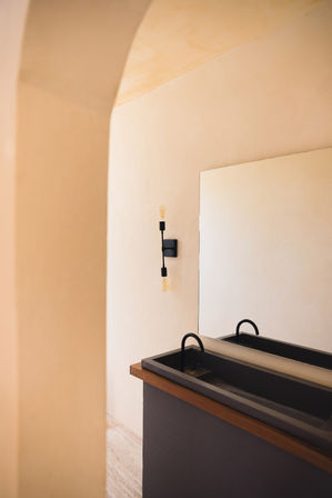 Sunlit minimalist bathroom interior in warm beige tones with an arched plaster doorway, large rectangular mirror, black double basin sink with curved faucets, wood-trimmed counter and exposed-bulb wall sconce.