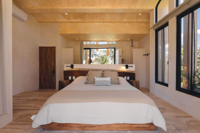 Bright minimalist tropical bedroom with a large bed dressed in neutral linens, wooden headboard, bedside sconces, vaulted ceiling and floor-to-ceiling windows framing palm trees and a relaxed beachy vibe.