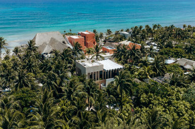 Aerial view of tropical beachfront villas and thatched-roof buildings tucked among palm trees along a turquoise ocean