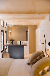 Sunlit neutral-toned bedroom with bed in the foreground and an open-plan bathroom featuring a wooden floating sink counter with black faucets, large mirror, woven basket and warm recessed and wall lighting.