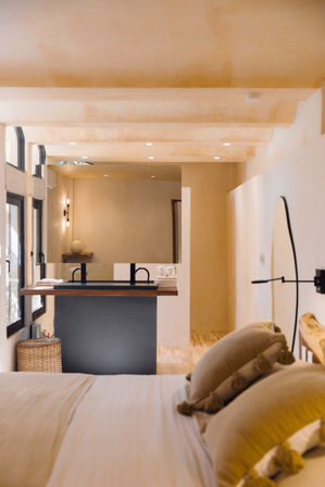 Sunlit neutral-toned bedroom with bed in the foreground and an open-plan bathroom featuring a wooden floating sink counter with black faucets, large mirror, woven basket and warm recessed and wall lighting.