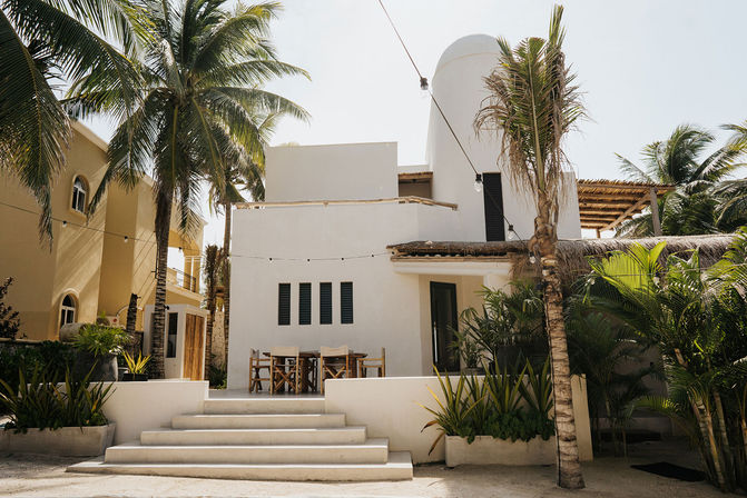 Sunlit coastal villa with white stucco façade, palm trees, sand steps and a wooden outdoor dining area under string lights.