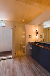 Sunlit minimalist bathroom with walk-in glass shower, travertine tile floor, wood-trimmed black vanity with double sinks, warm beige plaster walls and exposed-bulb sconces