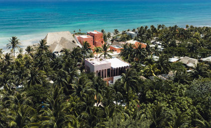 Aerial shot of colorful pink and terracotta seaside villas nestled in dense palm trees along a turquoise tropical coastline and sandy beach