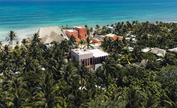 Aerial shot of colorful pink and terracotta seaside villas nestled in dense palm trees along a turquoise tropical coastline and sandy beach