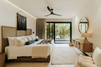 Sunlit modern tropical bedroom with king-size bed, woven rattan headboard, neutral linens and bolster pillows, black ceiling fan, sliding glass doors opening to lush greenery, wood console, round mirror and woven rug