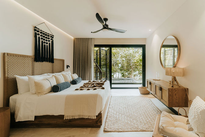 Sunlit modern tropical bedroom with king-size bed, woven rattan headboard, neutral linens and bolster pillows, black ceiling fan, sliding glass doors opening to lush greenery, wood console, round mirror and woven rug