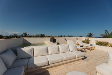 Sun-soaked coastal rooftop terrace with white cushioned sofa, two wooden sun loungers with striped towels, small plunge pool, tropical palms and ocean view under a clear blue sky