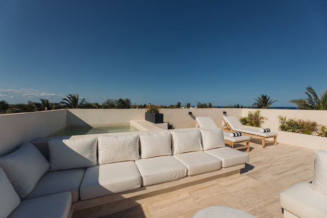 Sun-soaked coastal rooftop terrace with white cushioned sofa, two wooden sun loungers with striped towels, small plunge pool, tropical palms and ocean view under a clear blue sky