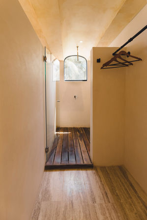 Sunlit minimalist walk-in shower with arched window, overhead rain shower, wooden slatted floor, smooth plaster walls and wooden hangers — tropical, resort-style vibe.