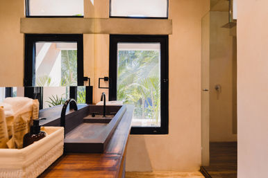 Sunny tropical bathroom with a long wooden trough sink and matte black faucets, towel and soap basket, windows framing palm trees and a glass walk-in shower.