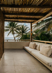 Tropical beachfront balcony with woven wooden pergola, long cushioned outdoor bench, swaying palm trees and a calm ocean view
