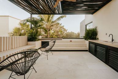 Modern tropical rooftop terrace with striped built-in bench, black wire chairs, outdoor kitchenette and sink, shaded wooden pergola and palm trees in the background