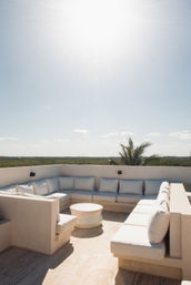 Sun‑drenched rooftop terrace with L-shaped beige cushioned sofa and round coffee table, low stucco walls overlooking green treeline and a palm under a clear blue sky.