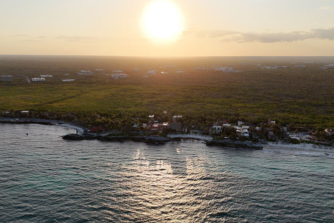 Aerial sunset view of a tropical coastline with beachfront homes and palm trees, golden sun reflecting on rippling ocean waves and a narrow sandy shore.