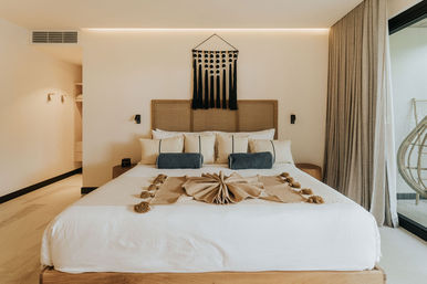 Sunlit modern bedroom with king bed, woven rattan headboard, neutral linens and layered pillows with navy bolsters, macramé wall hanging, tasseled beige throw, floor-to-ceiling curtains and glass door opening to a balcony with a hanging chair.