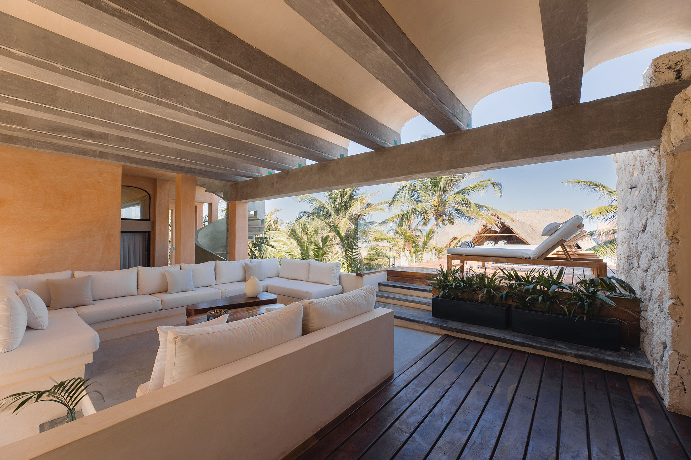 Sun-soaked beachfront lounge at a tropical resort with a large white sectional sofa, wooden deck, raised sunbeds, palm trees and ocean view.