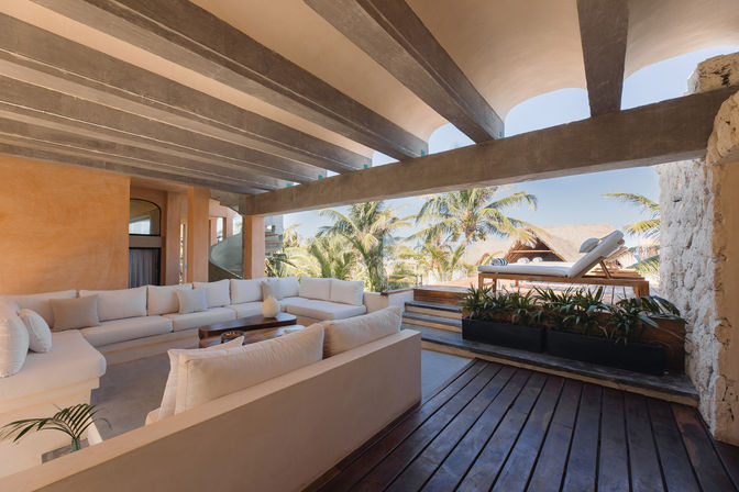 Sun-soaked beachfront lounge at a tropical resort with a large white sectional sofa, wooden deck, raised sunbeds, palm trees and ocean view.