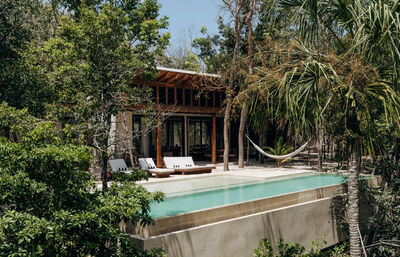 Modern tropical villa with elevated infinity pool and wooden deck, three white lounge chairs and a hammock strung between trees, surrounded by dense palm and jungle greenery.