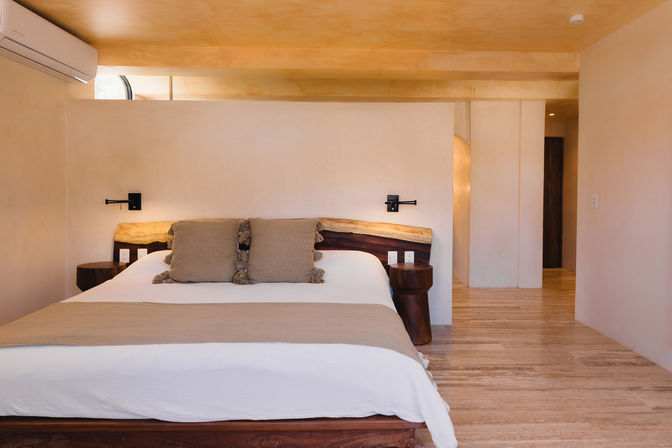 Sunlit minimalist bedroom with large bed, live-edge wooden headboard, neutral linens and tassel pillows, wall-mounted reading lamps, round wooden side tables, and wall air conditioner