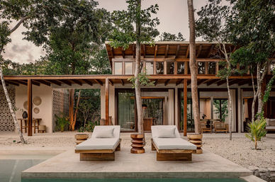 Modern tropical villa pool deck with two cushioned wooden loungers, sculptural side tables, surrounding trees and open-air wood-beam architecture.