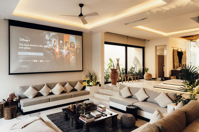 Modern open-plan living room with a large wall projector screen showing a movie image, wide built-in grey sofas with neutral throw pillows, low dark wood coffee table stacked with books, potted plants and terracotta vase, stepped seating area and floor-to-ceiling windows — bright, neutral-toned space ideal for movie nights.