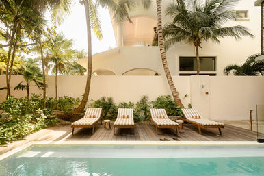 Sunlit tropical pool deck with four striped lounge chairs on wooden decking, palm trees and white villa facade