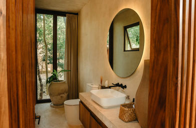 Sunlit modern rustic bathroom with round backlit mirror, white vessel sink on a stone countertop, black wall faucet, woven basket and large potted plant by a floor-to-ceiling window overlooking green trees.