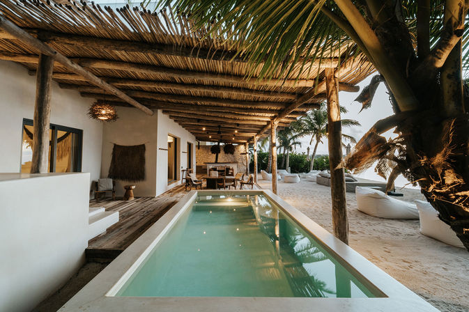 Tropical beachfront villa patio with a narrow private plunge pool under a rustic thatched pergola, wooden dining table, palm trees and sandy lounge area with beanbag seats at golden hour.