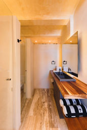 Warm, minimalist resort-style bathroom with beige plaster walls and ceiling, travertine floor, long wooden vanity with black double trough sink and matte black faucets, large mirror, and striped towels with palm motif.