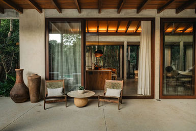 Modern tropical villa patio with two rustic wooden lounge chairs and white cushions around a round stone coffee table with a succulent, large terracotta vases, sliding glass doors and warm-lit interior with exposed wooden ceiling beams.