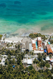 Aerial view of a tropical coastline with turquoise ocean and sandy beach lined by palm trees, thatched huts and colorful beachfront buildings with swimming pools.