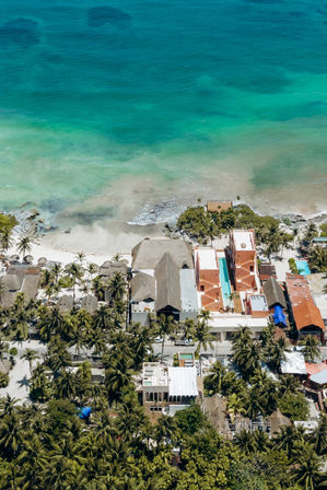Aerial view of a tropical coastline with turquoise ocean and sandy beach lined by palm trees, thatched huts and colorful beachfront buildings with swimming pools.