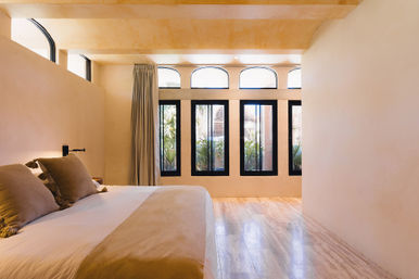 Sunlit minimalist bedroom with low bed, beige linens and throw pillows, arched transom windows above tall black-framed panes overlooking tropical plants, light wood floor and warm neutral walls.