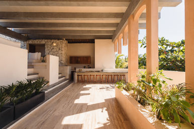 Sunlit tropical terrace with an open-air kitchen, long counter lined with wooden bar stools, stone accent wall, arched colonnade and lush planter boxes.