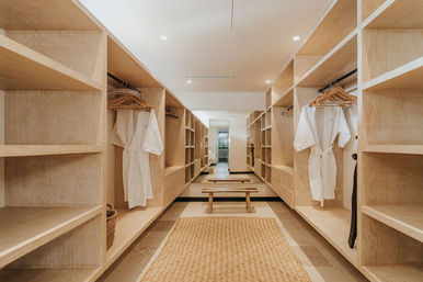 Bright modern walk-in closet with light oak built-in shelving, hanging white spa robes, woven jute rug and wooden bench facing a full-length mirror.