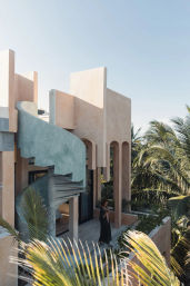 Sunlit pastel villa with a mint-green spiral concrete staircase, arched terrace framed by palm trees, and a lone person on the balcony enjoying a drink under a clear blue tropical sky.
