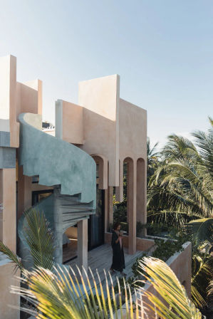 Sunlit pastel villa with a mint-green spiral concrete staircase, arched terrace framed by palm trees, and a lone person on the balcony enjoying a drink under a clear blue tropical sky.