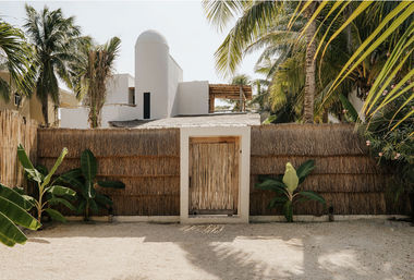 Rustic bamboo gate set in a thatched fence leading to a white stucco coastal villa, sandy courtyard framed by palm trees and banana plants in a tropical setting