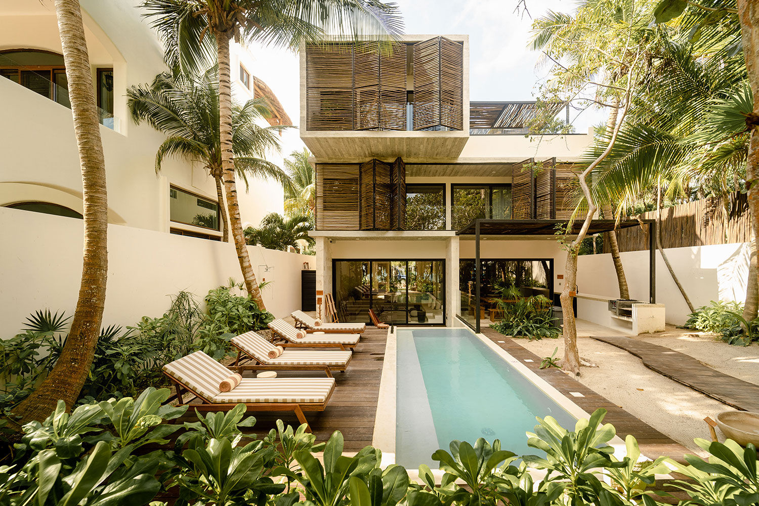 Sunny tropical modern villa courtyard with a narrow lap pool, wooden deck lined with striped lounge chairs, palm trees, lush greenery and two-story balcony with wooden privacy screens