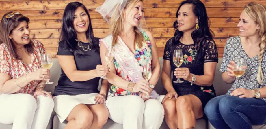 Five women seated on a couch against a wooden wall, toasting with white wine while celebrating a bride-to-be wearing a veil and sash at a bachelorette party.