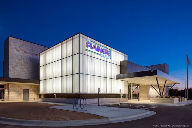 Glowing modern glass-cube building exterior at dusk with frosted-panel facade, covered entrance canopy, bike rack and flagpole on a clean paved forecourt.