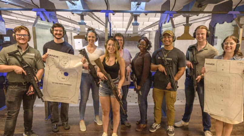 Group of nine people in an indoor shooting range lane wearing ear protection and safety glasses, holding rifles and paper targets with bullet holes after target practice.