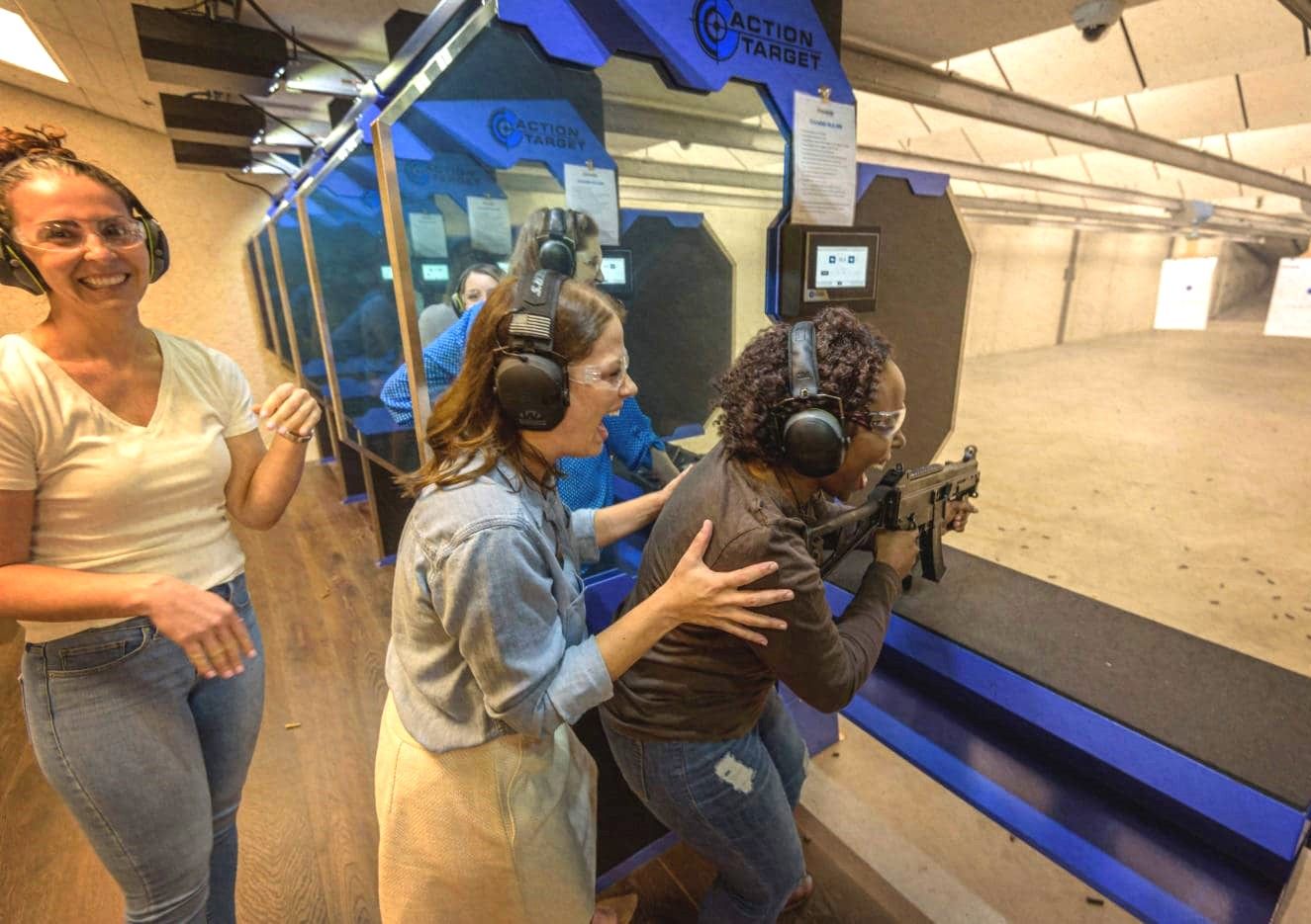 Three women at an indoor shooting range; one aims a rifle at distant paper targets while others assist and cheer, all wearing ear protection and safety glasses.