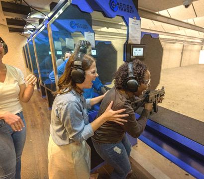 Three women at an indoor shooting range; one aims a rifle at distant paper targets while others assist and cheer, all wearing ear protection and safety glasses.
