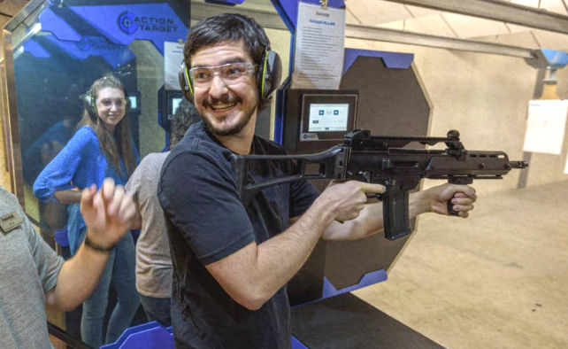 Smiling person wearing safety glasses and earmuffs holds a rifle at an indoor shooting range lane while others watch from adjacent booths.