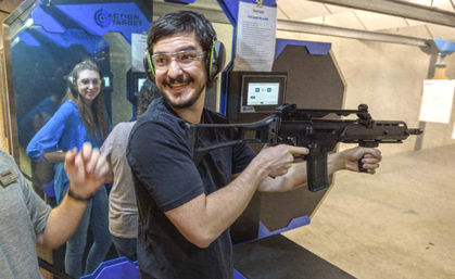 Smiling person wearing safety glasses and earmuffs holds a rifle at an indoor shooting range lane while others watch from adjacent booths.