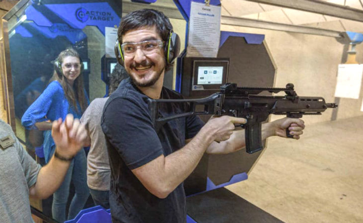 Smiling person wearing safety glasses and earmuffs holds a rifle at an indoor shooting range lane while others watch from adjacent booths.